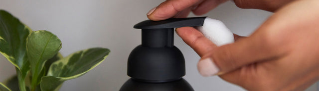 A close-up of a hand dispensing Saje foaming hand soap with a plant in the background