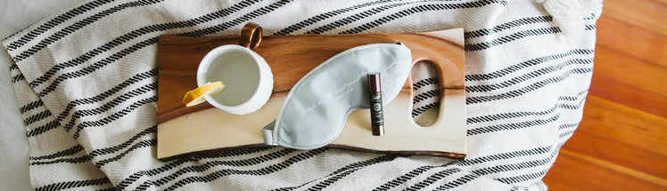 Saje Sleep Well Roll-on, eye mask, and cup of lemon water displayed on a tray on top of fresh bed sheets