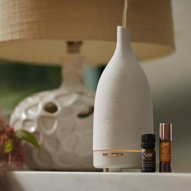 Aroma Om Diffuser in white, Liquid Sunshine diffuser blend and Peppermint Halo Roll-On sitting on a white surface  next to a lamp.