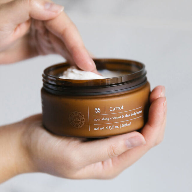 A right hand holding an open Carrot Body Butter container while the left hand scoops out some body butter
