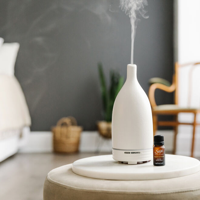 A white diffuser diffusing liquid sunshine in a grey room with a chair, basket and bed.