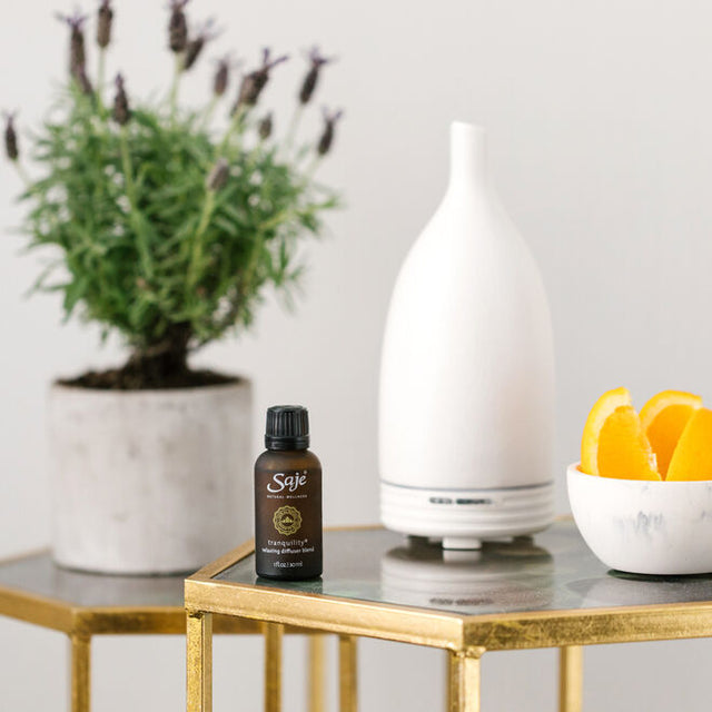 Tranquility 1 fl oz relaxing diffuser blend in bottle next to an Aroma Om white with a bowl of orange slices and a lavender plant in the back