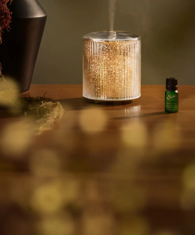 The Aroma Lustre diffuser rests, running, on a wood table beside a holiday diffuser blend and a decorative vase.