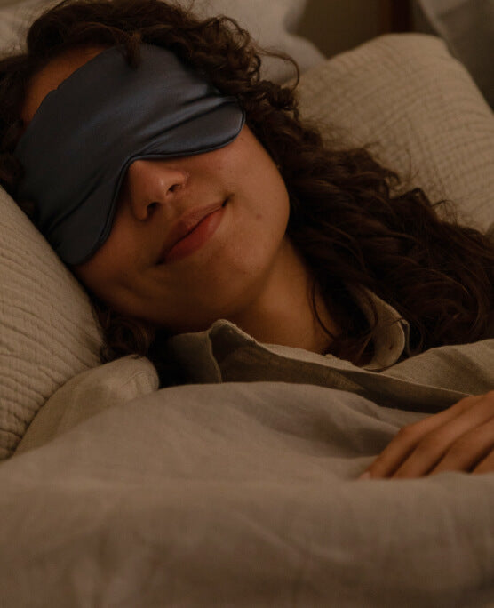A young woman is asleep in her bed. She wears a silky blue eye mask.