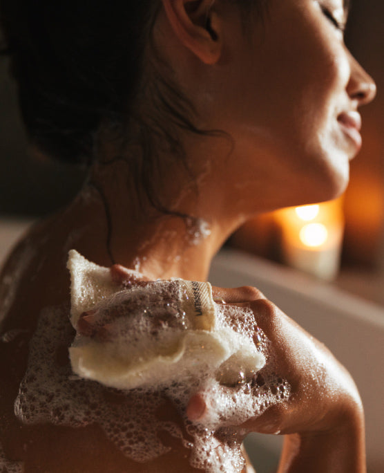 A smiling person with eyes closed in the bath lathers up her shoulder with soap.