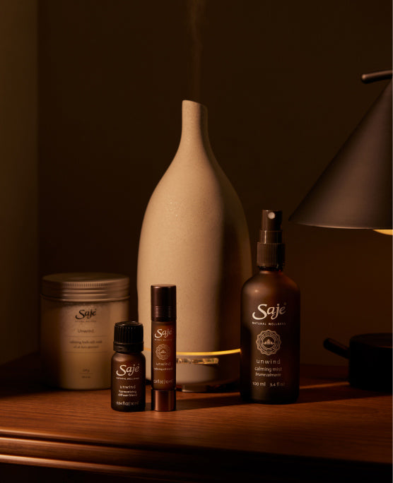 Dimly-lit Saje products—including a diffuser, diffuser blends, bath salts, and roll-ons—rest atop a wooden shelf.