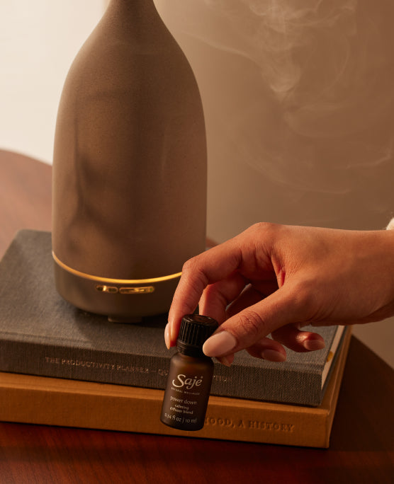 A hand holding a brown bottle of Power Down Diffuser Blend with a diffuser behind.