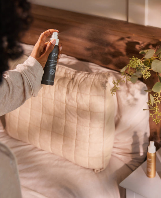 A person spritzes a Saje product on their beige pillowcase and linens.