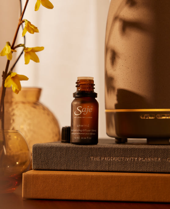 A brown bottle of Unwind Diffuser Blend sits atop two books with yellow flowers and a diffuser in the background.