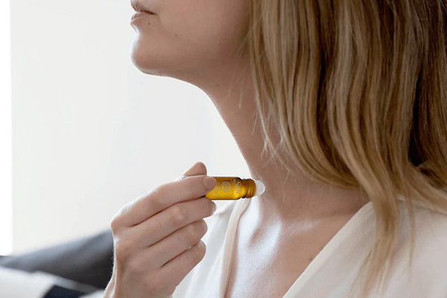 A woman raises her arm, holding a Saje remedy roll-on from her bedsheets.