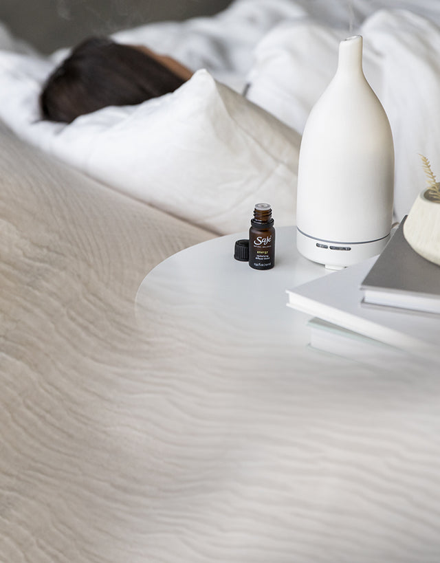 A woman sleeping with a white Saje Aroma Om diffuser and diffuser blend on her bedroom side table.