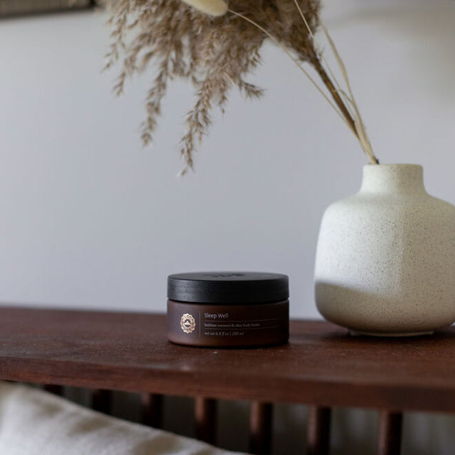 Sleep Well Body Butter beside a vase with dried flowers on a wooden headboard