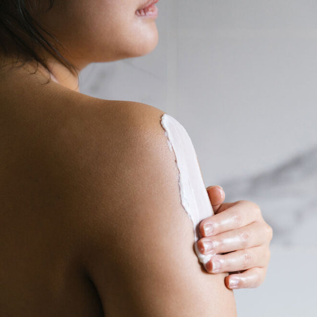 A woman applying body butter to her bare shoulder