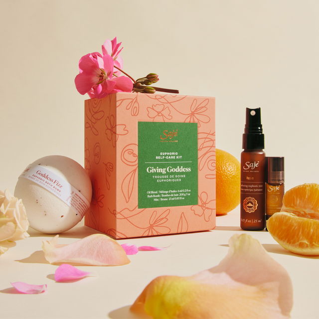 A limited-edition Giving Goddess wellness kit—bath bomb, face mist, roll-on—along with pink flowers and fresh citrus.