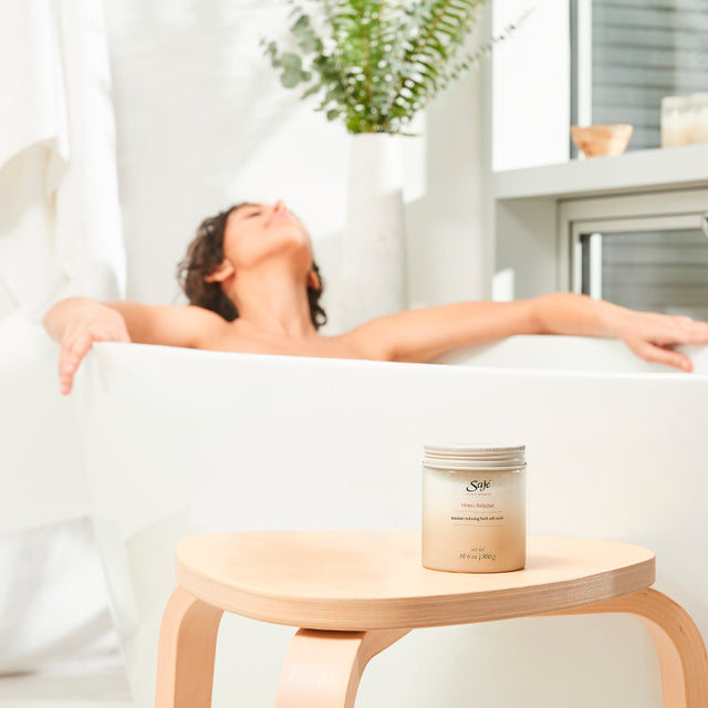 Person relaxing in the bath tub with Saje bath salts on a stool in the forefront